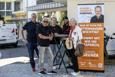 Daniel Gruner im Gespräch mit Dettenhausen Daniel Gruner und mehrere Besucher unterhalten sich an einem Stehtisch vor seinem Wahlkampfaufsteller.