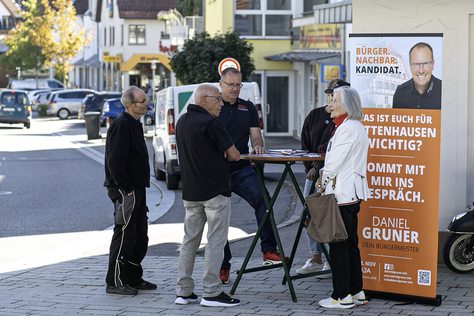 Offener Austausch auf dem Parkplatz La Banca Mehrere Besucher unterhalten sich am Infostand von Daniel Gruner neben seinem Wahlkampfaufsteller.